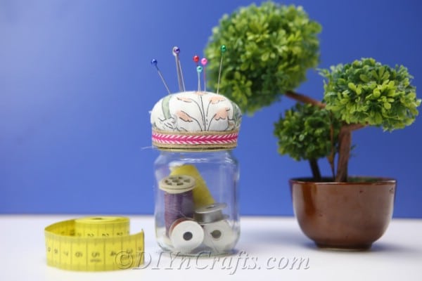 A completed pincushion jar with a small plant and a tape measure against a blue background.