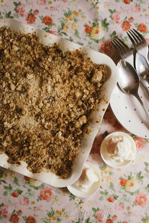 Overhead picture of a prepared apple crumble recipe in baking dish sitting on floral tablecloth next to serving dishes