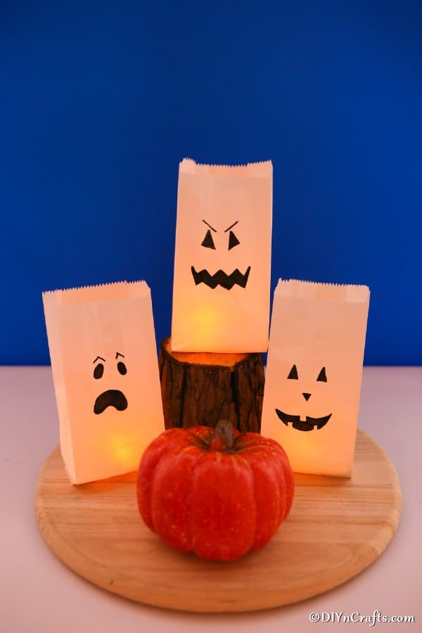 Luminary bags decorated as ghosts sitting on a round wooden plate in front of a blue background