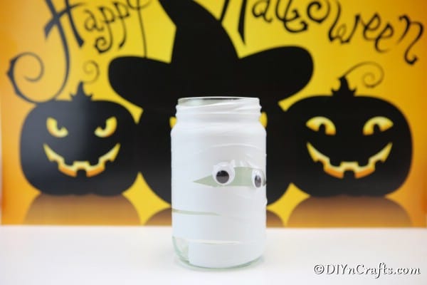 Mason jar lights mummy sitting on a white counter with yellow halloween banner behind it