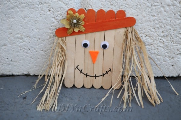 Completed scarecrow from how to make a scarecrow out of craft sticks tutorial sitting against a white wall