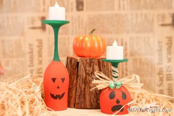 Two wine glass pumpkin art pieces sitting on a tan surface with a block of wood and mini pumpkins in the background