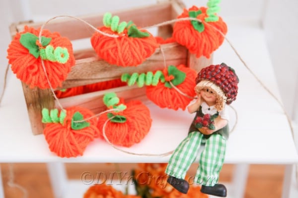 A diy fall decor yarn pumpkin garland displayed on a white shelf with a scarecrow doll
