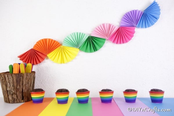 Beautiful rainbow paper fan garland on a white wall behind cupcakes on table