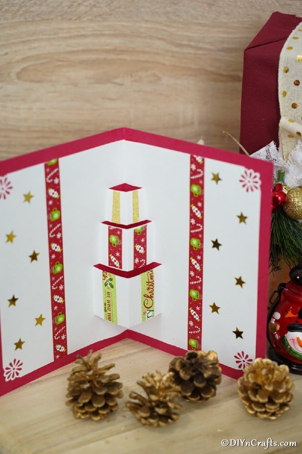 A holiday gift box 3D christmas card displayed next to pine cones and a wooden background