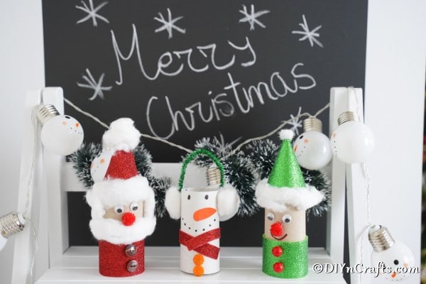 Three toilet paper roll crafts for Christmas displayed in front of a holiday chalkboard