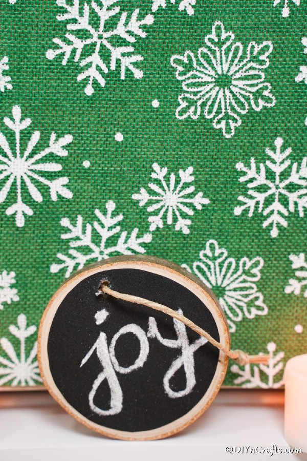 A wood slice chalkboard ornament leaning against a green snowflake background