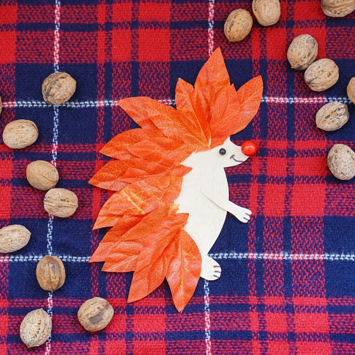 Red and blue plaid fabric with paper and leaf hedgehog on top next to pecans