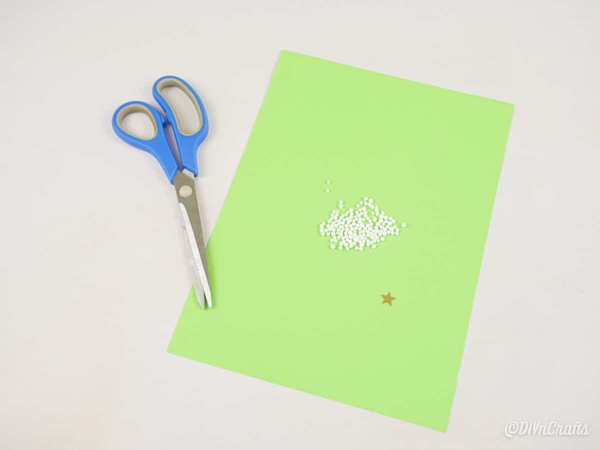 green paper on white table with blue scissors and white beads on top