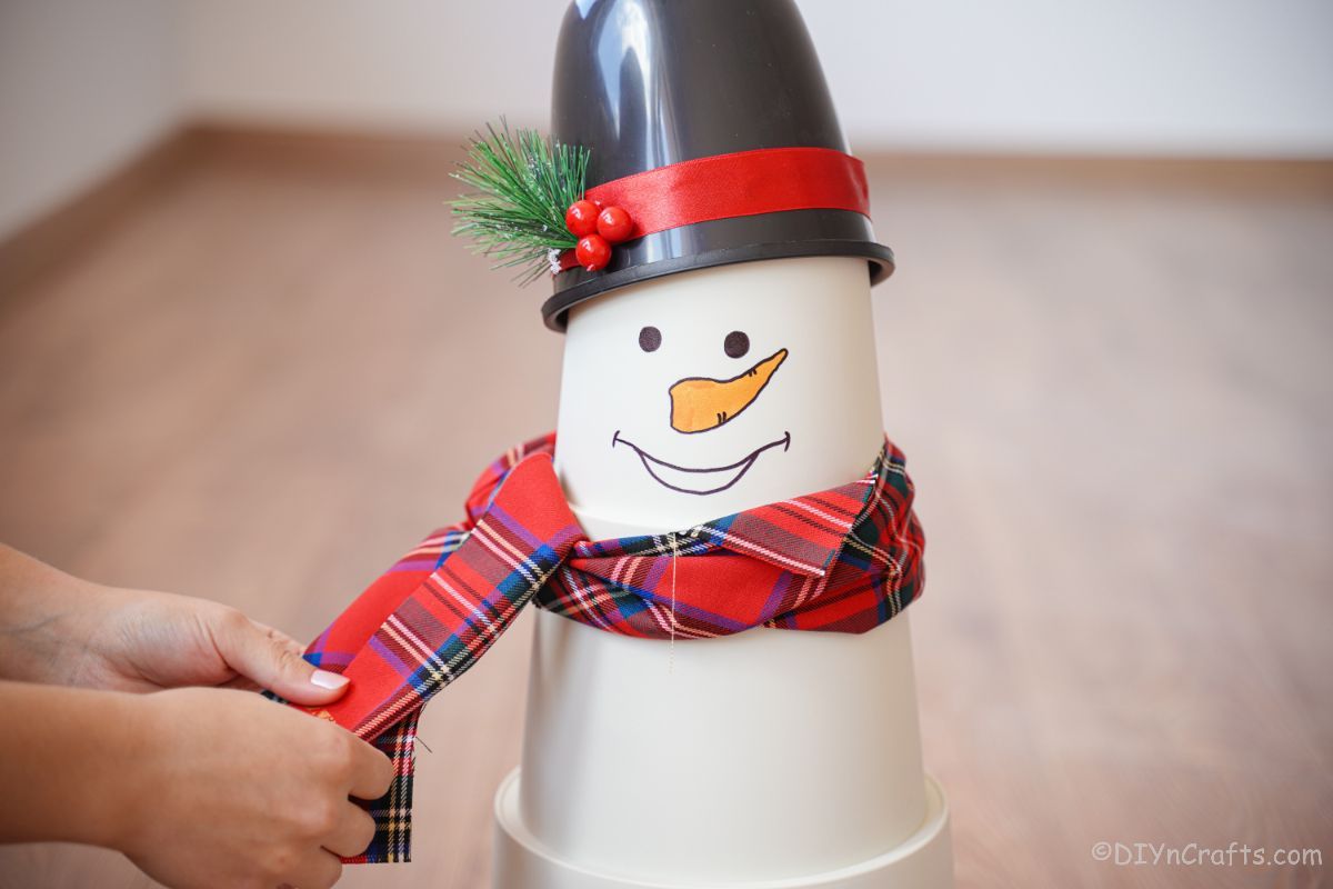 hands tying plaid scarf around neck of white flowerpot snowman