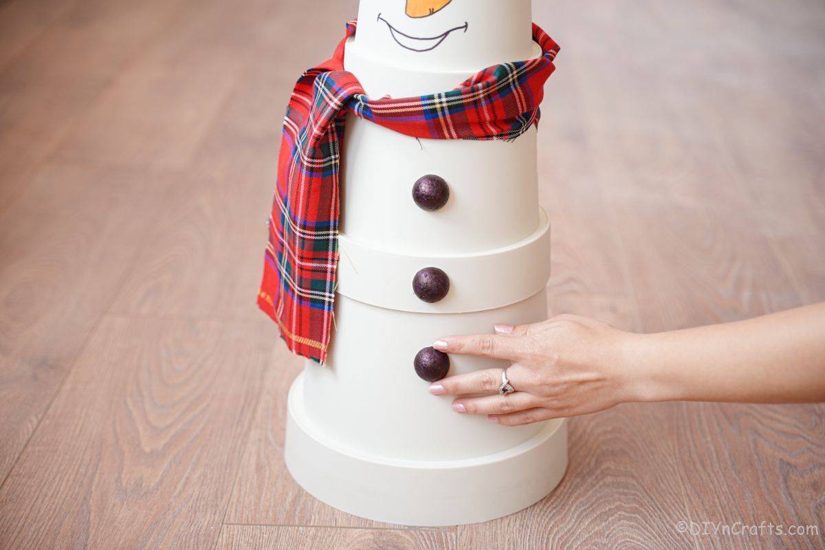 hand holding black button onto front of white flower pot snowman