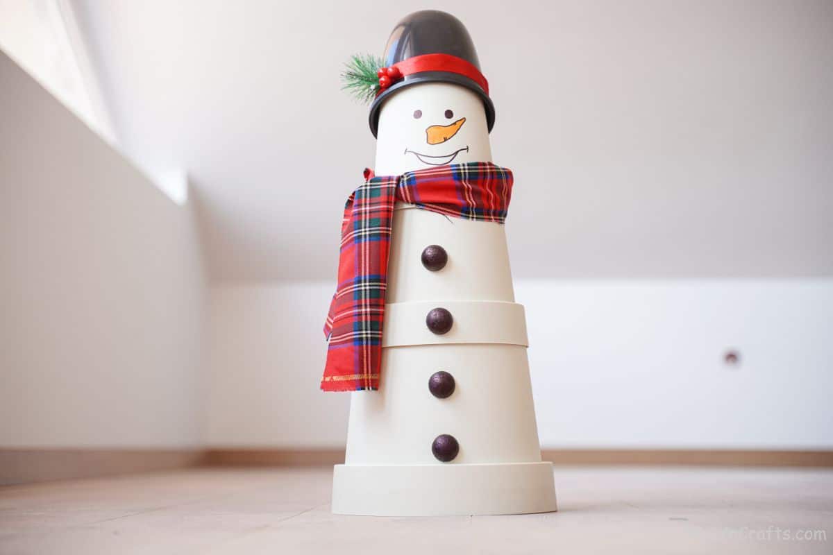 flowerpot snowman with black hat and plaid scarf on floor with white walls in background