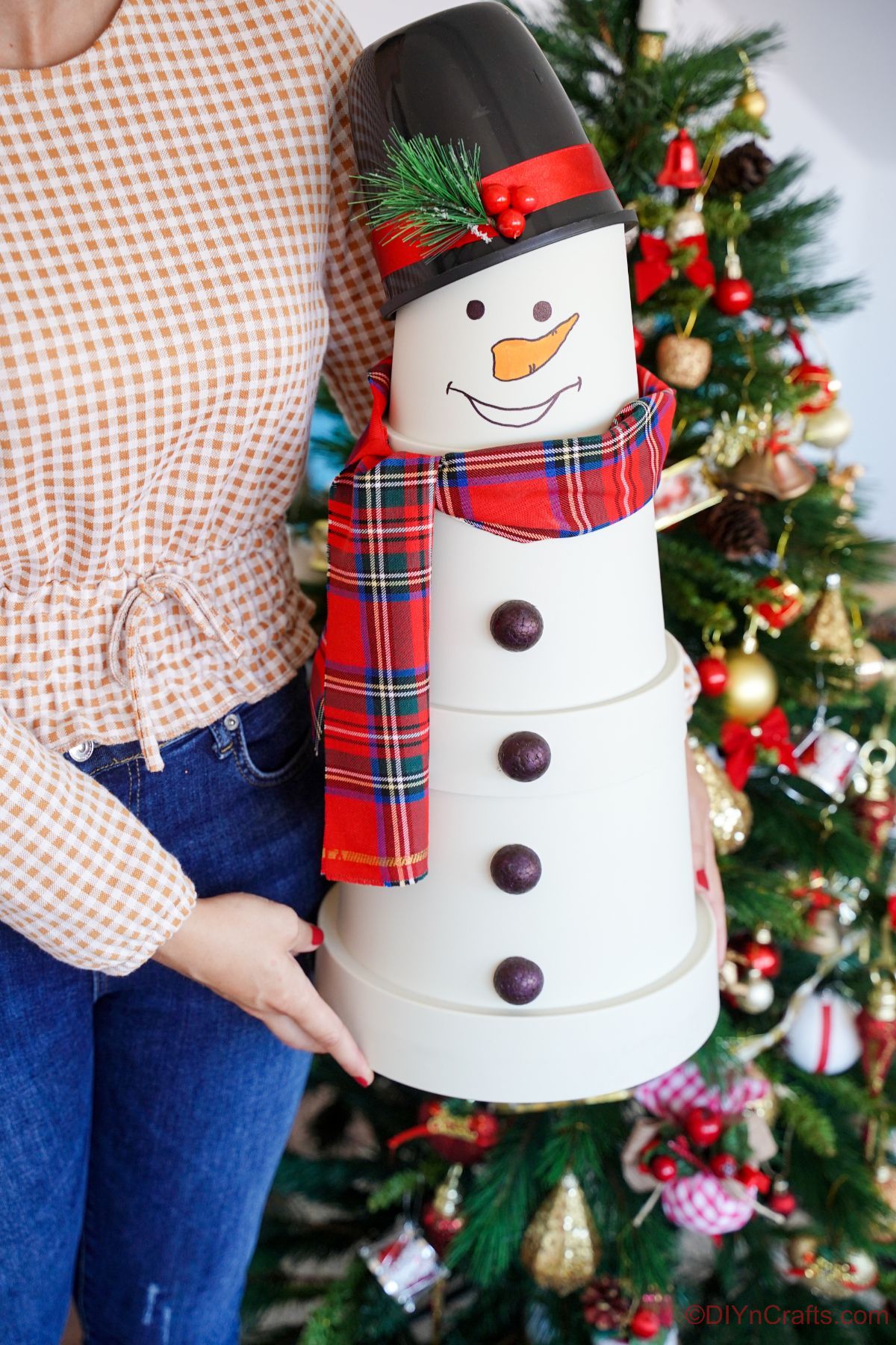 woman in jeans and peach shirt holding white clay pot snowman with red plaid scarf