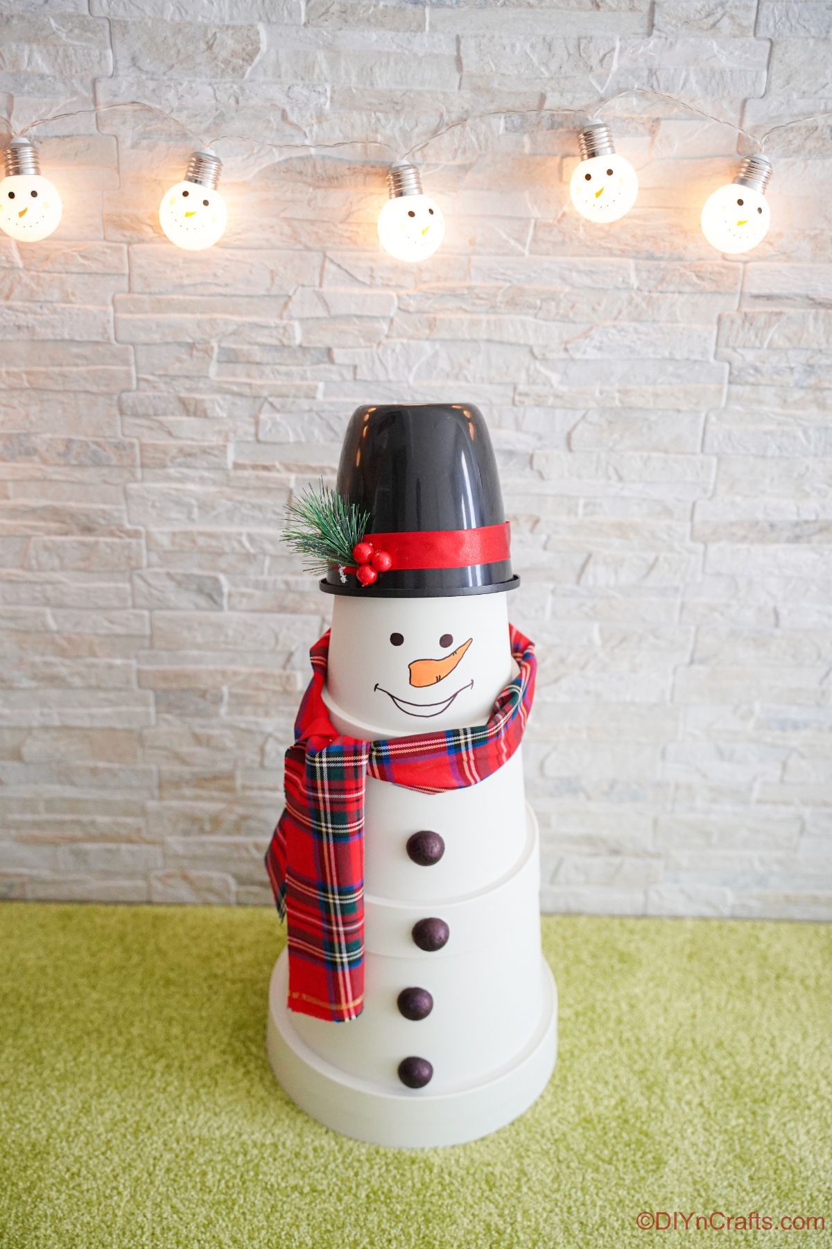 fake snowman on green floor with white brick background and lights hanging against wall