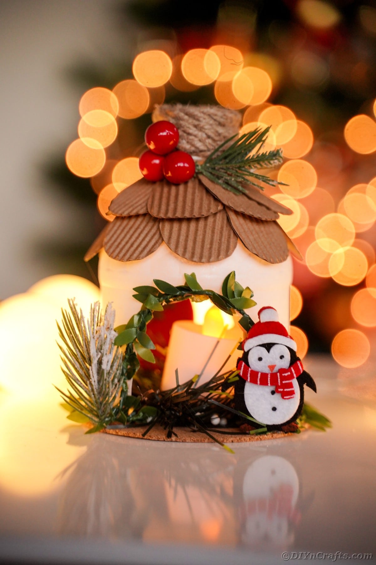 mini house with penguin on side and cardboard shingles sitting on table in front of twinkling holiday lights