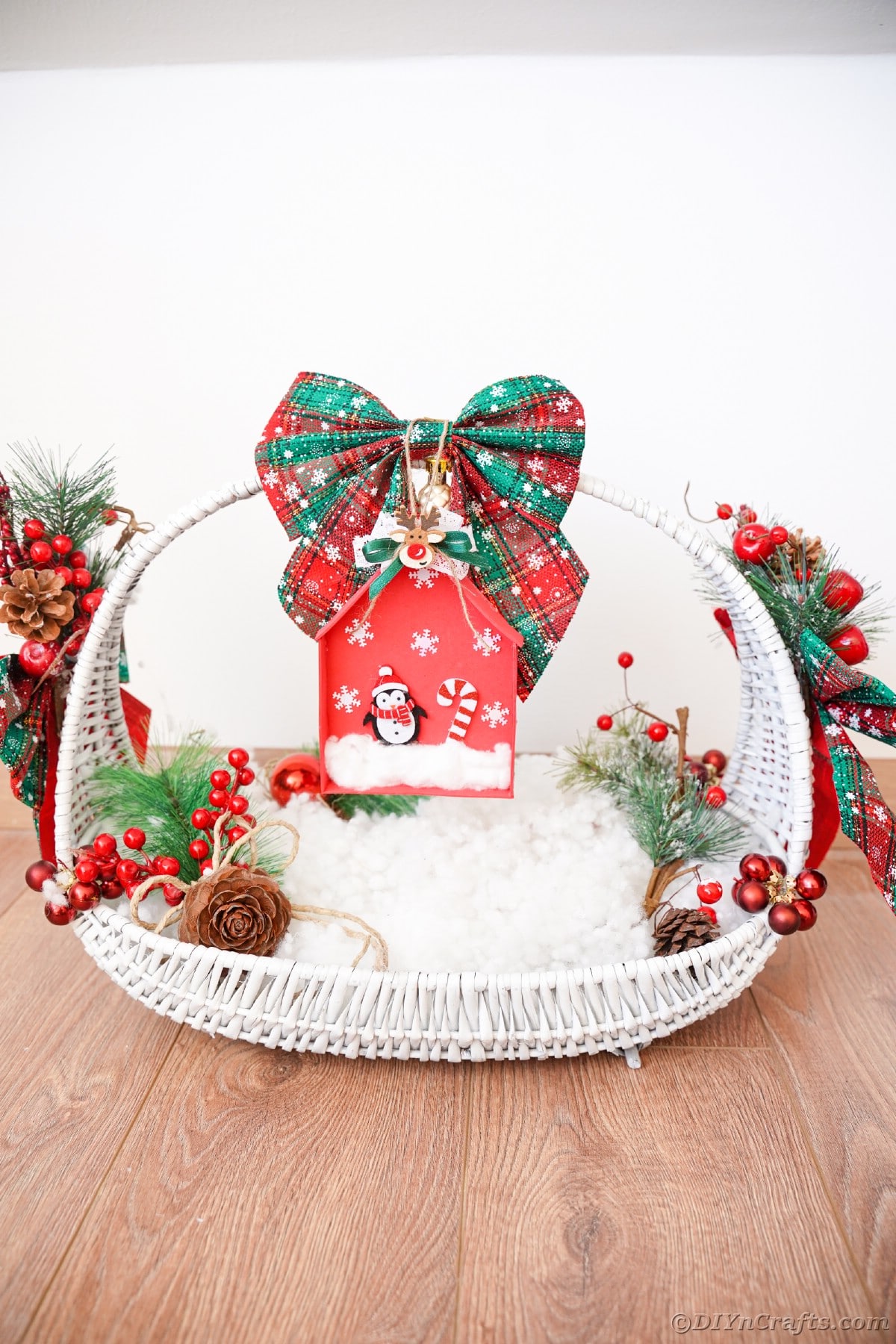 white basket on table filled with fake snow and a foam house ornament hanging beneath green bow