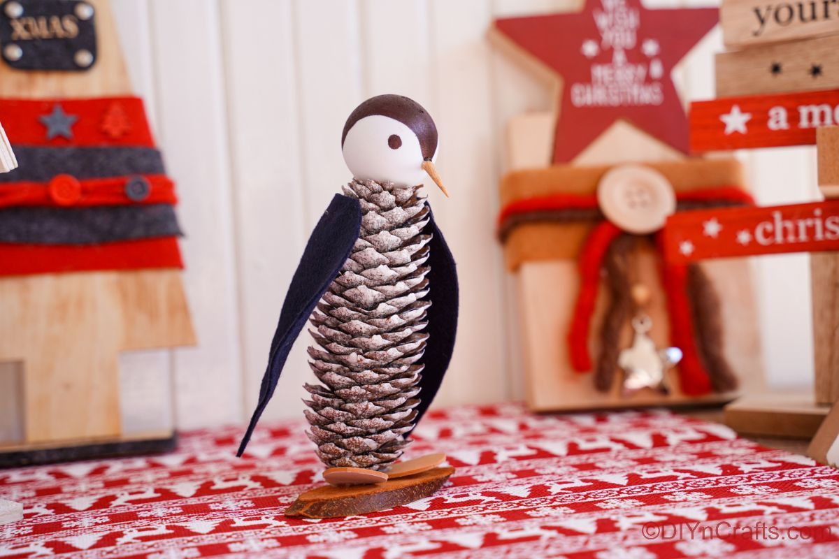 pinecone penguin on red and white paper with holiday decorations in background