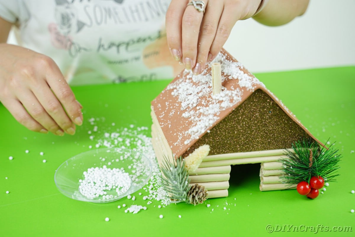 hands sprinkling fake snow on roof of miniature house