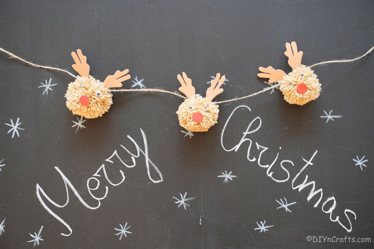 three pom pom reindeer on twine hanging over chalkboard with merry christmas message