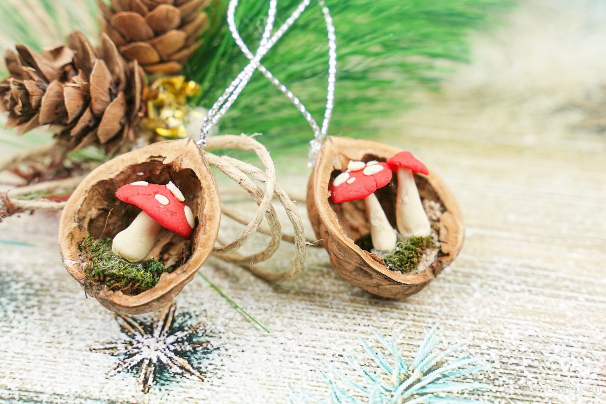 two walnut shell ornaments with red and white mushrooms laying on holiday theme paper by pinecones