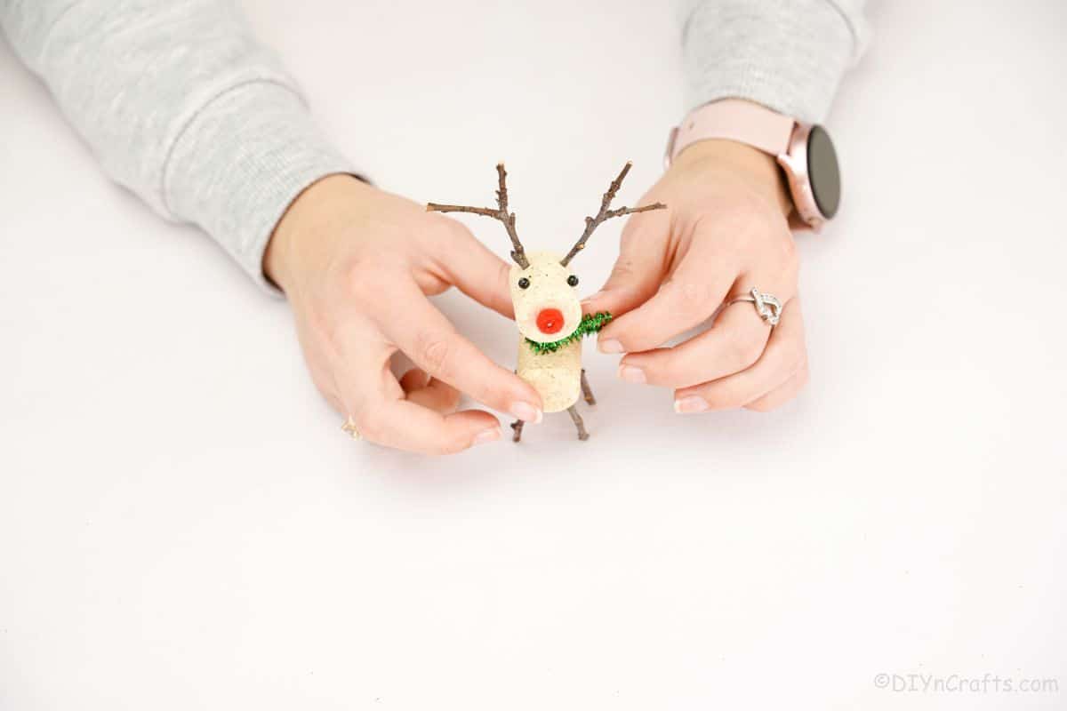 hand adding green pipe cleaner scarf to reindeer made of cork