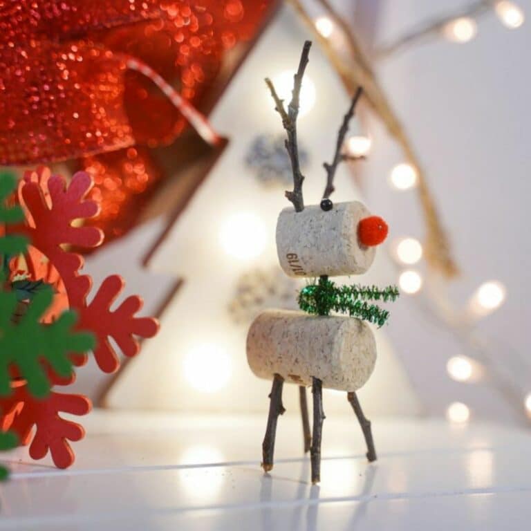 wine cork reindeer with green pipe cleaner scarf on table by red ribbon