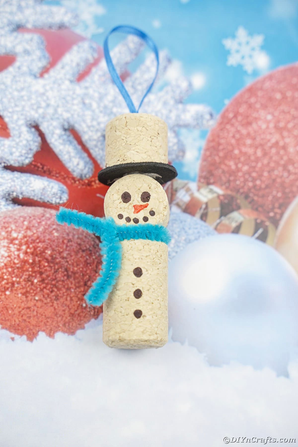 snowman ornament made of cork with blue pipe cleaner scarf on blue and white paper