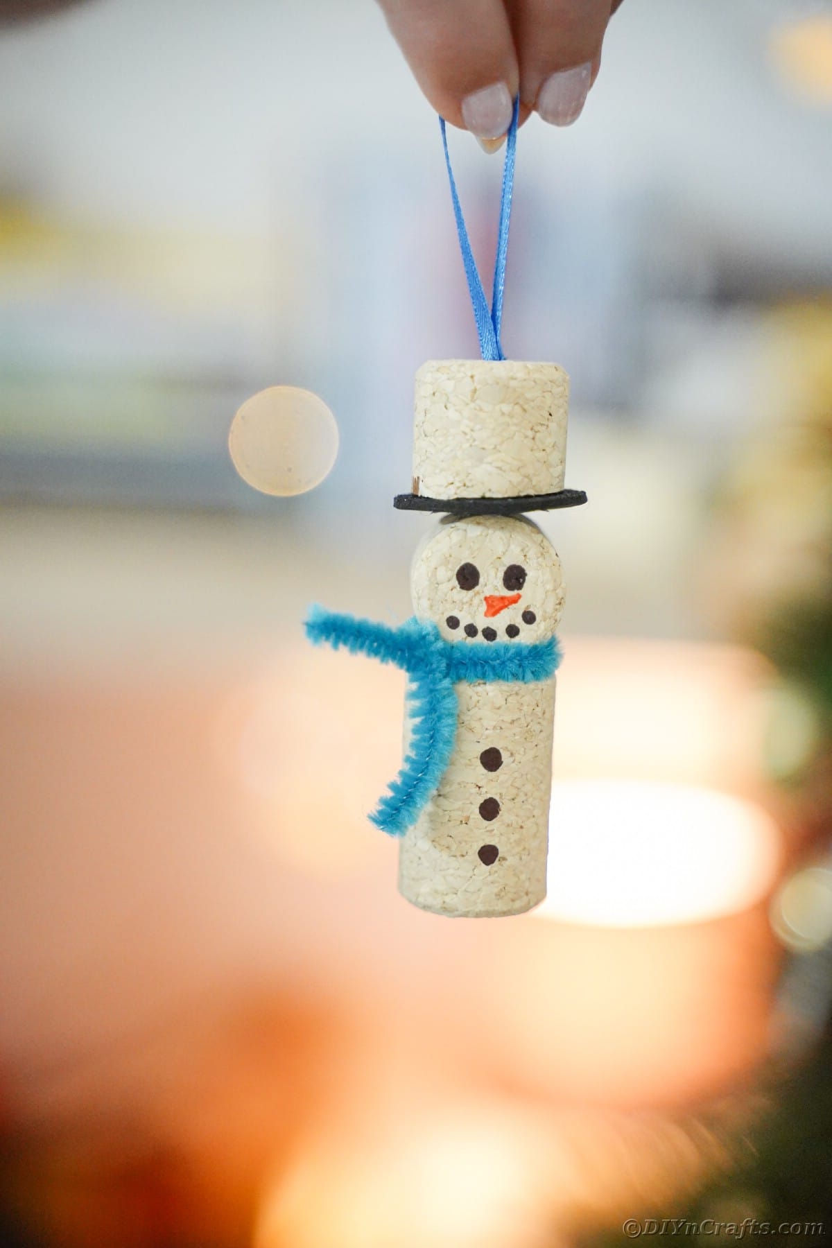 cork snowman with blue scarf being held in front of twinkling lights