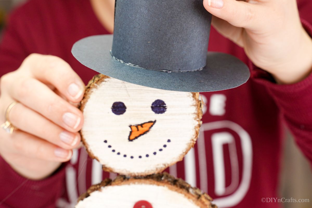 hand holding paper hat on top of wood slice snowman