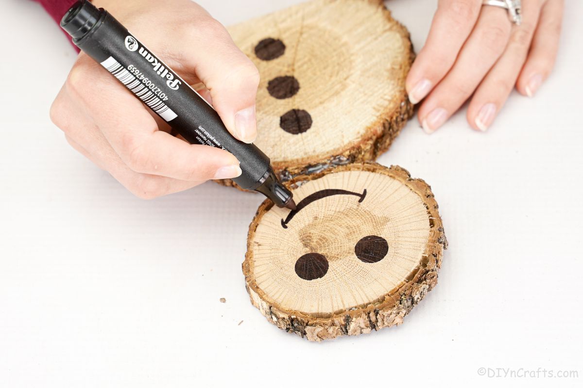 hand holding a black marker smiling at wooden slice on snowman's face