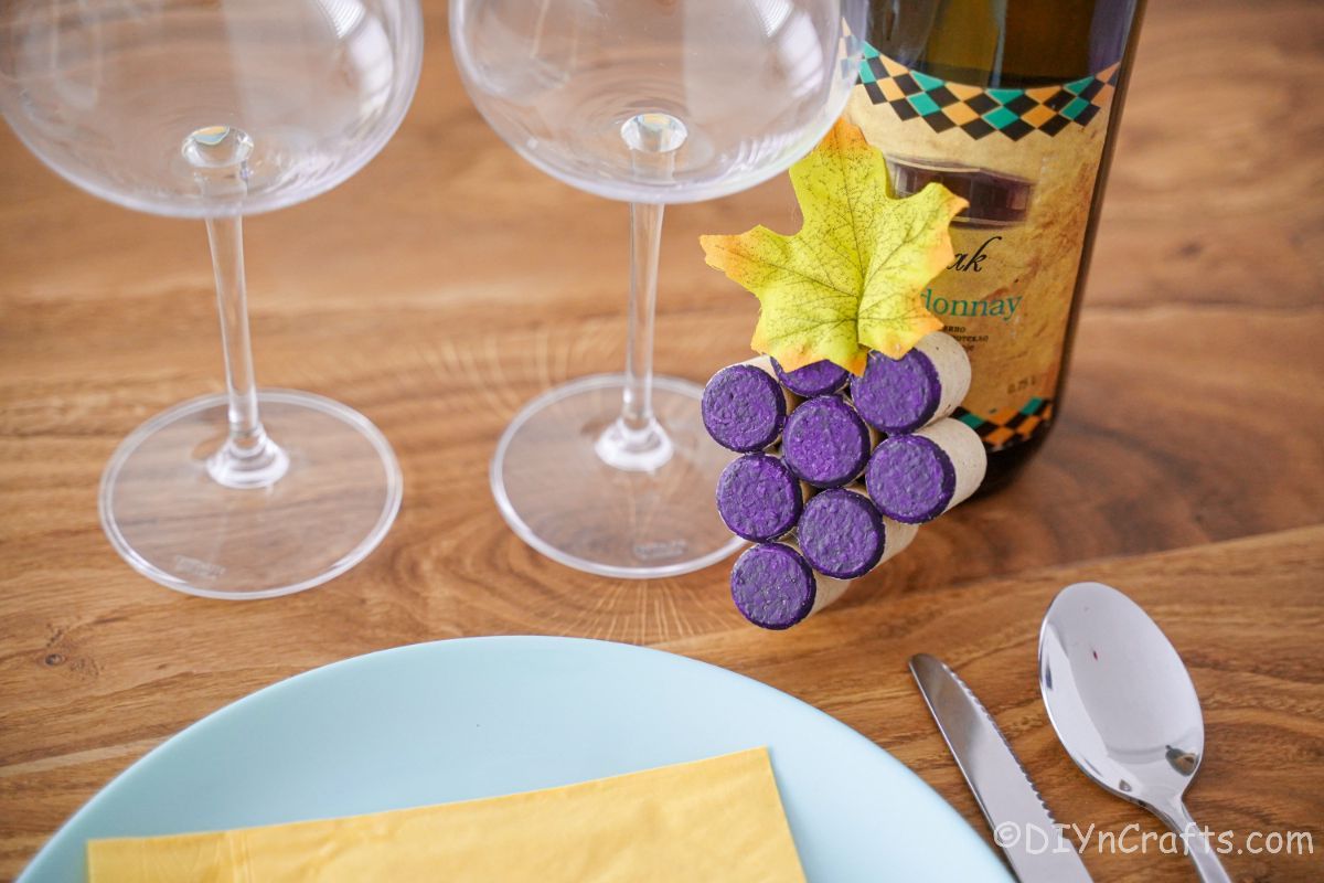 place setting with two wine glasses next to bottle of wine with cork grape cluster against bottle