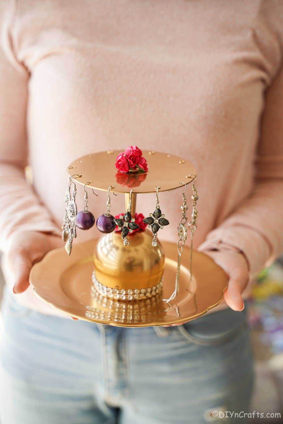 woman in jeans and pink sweater holding gold 2-tier jewelry stand