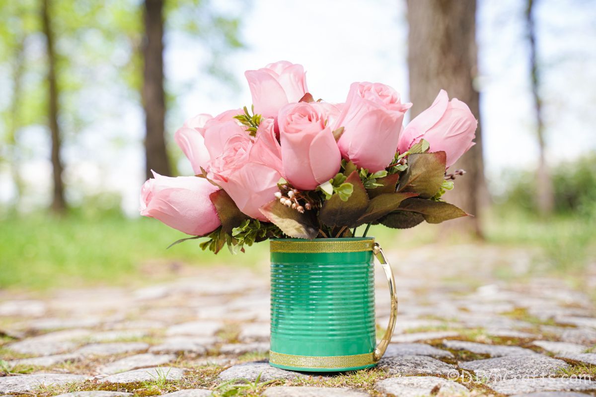 light green and gold tin can with gold handle on cobblestone path with pink roses