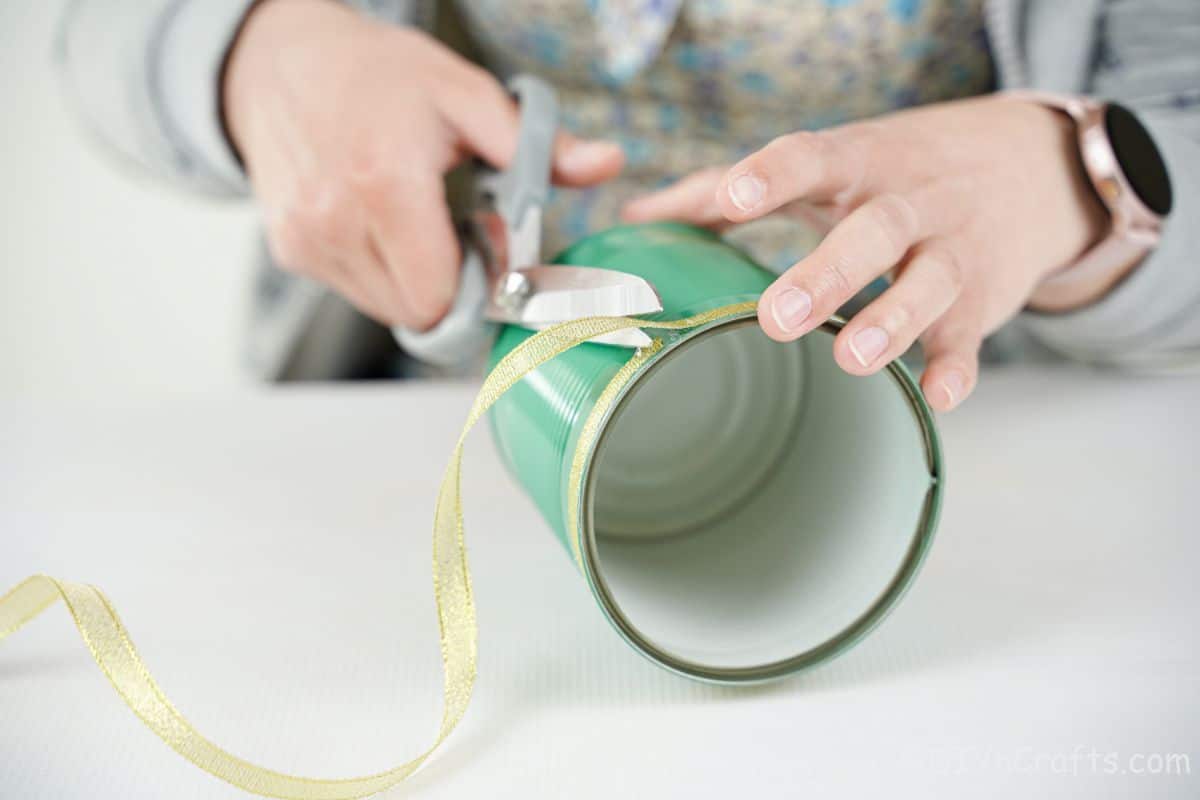 hand holding grey scissors to trim gold ribbon on green tin can