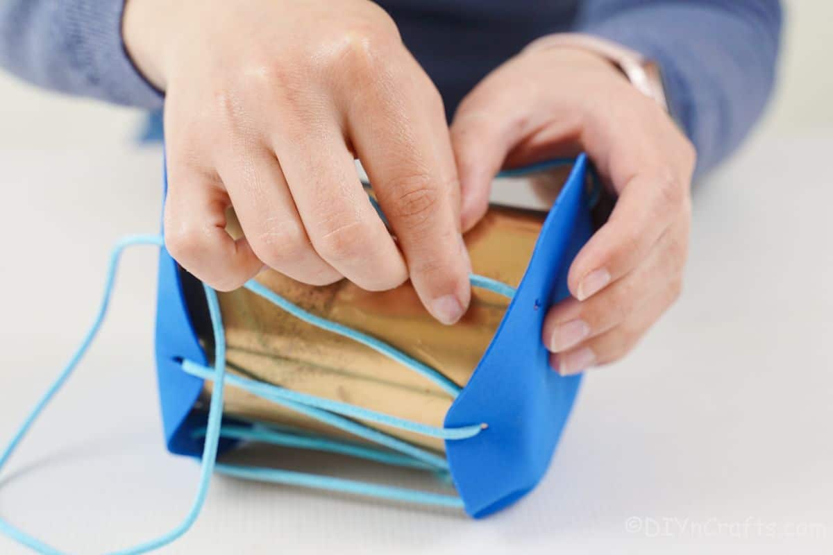 hands threading blue leather ribbon through holes on blue foam on top of gold tin can