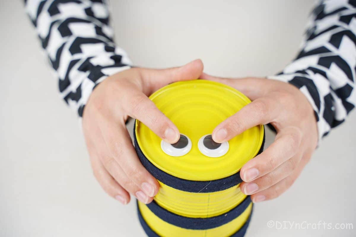 hand holding googly eyes to top of yellow tin can