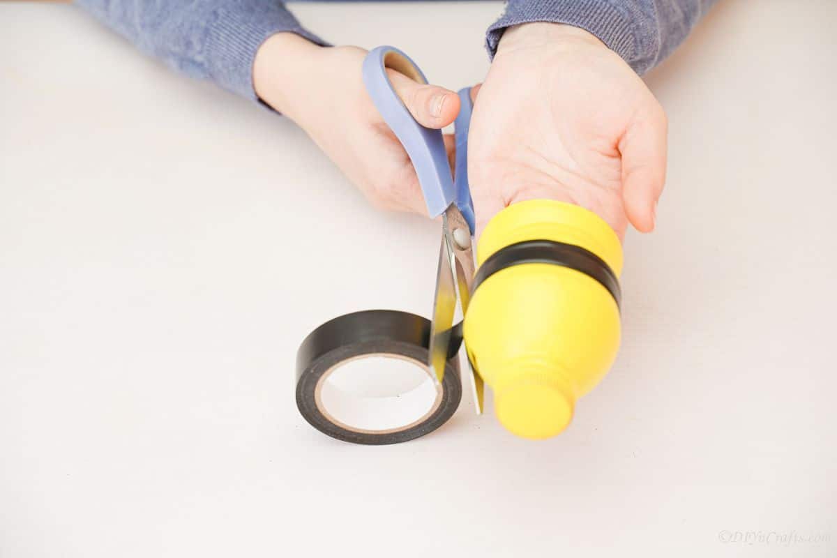 blue scissors cutting black tape on edge of yellow bottle