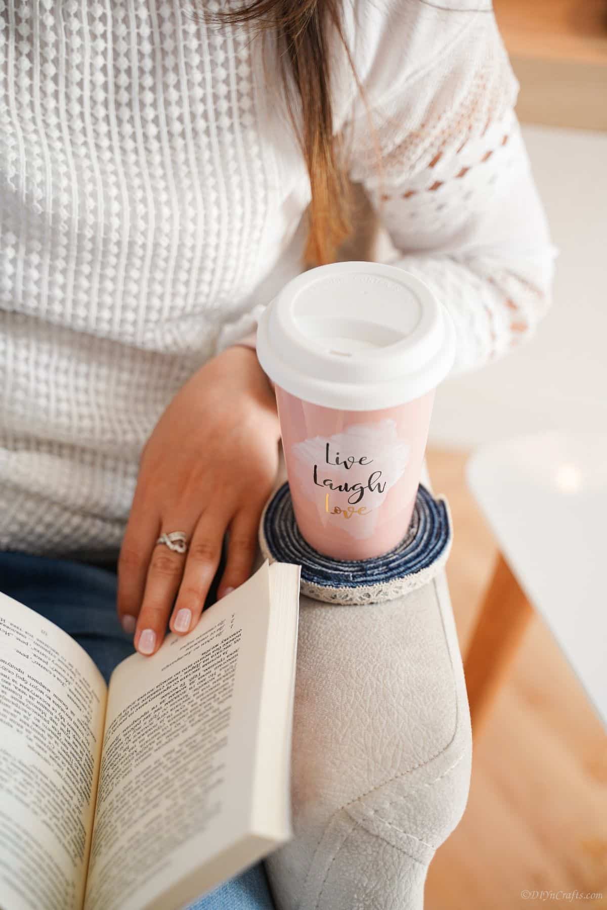 lady in white sweater in white chair with book in lap and coaster holding travel mug on arm of chair