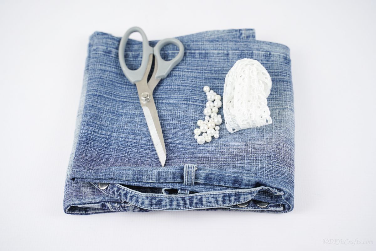 folded blue jeans on white table with gray scissors and white beads and lace