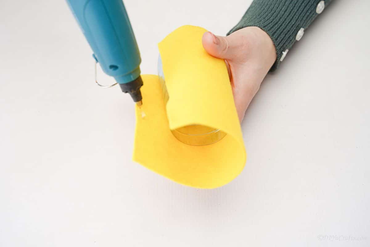 hand holding blue glue gun to attach yellow felt around bottle