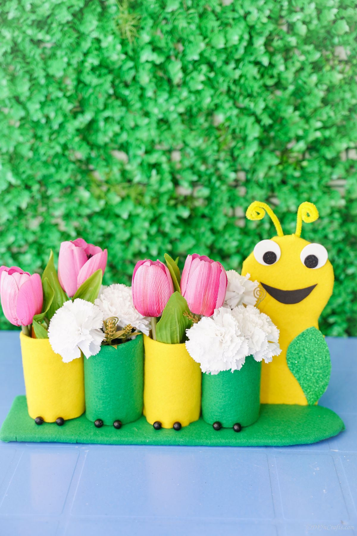 green and yellow caterpillar holding pink and white flowers in front of green shrub background