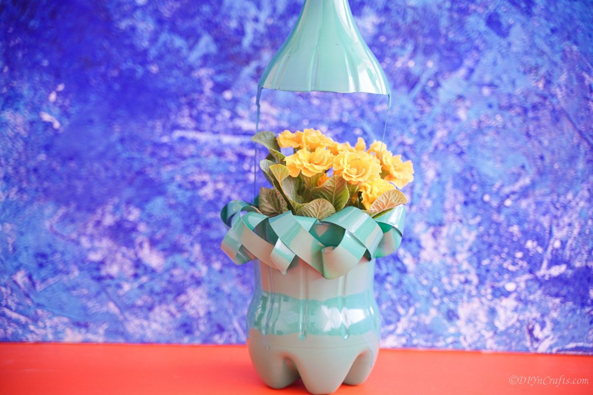 green upcycled bottle planter with yellow flowers on orange mat with blue background