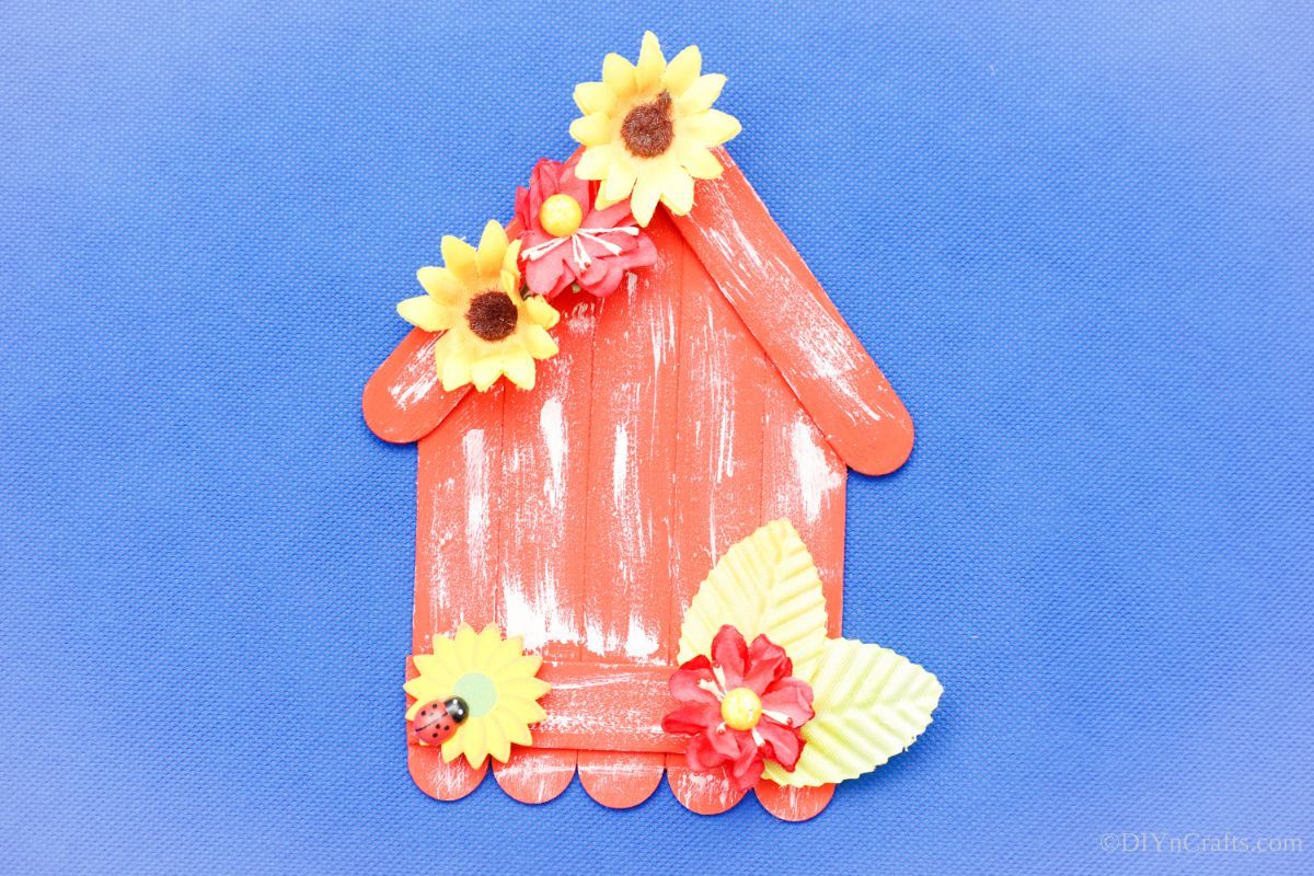blue background behind craft stick house with yellow flower accent